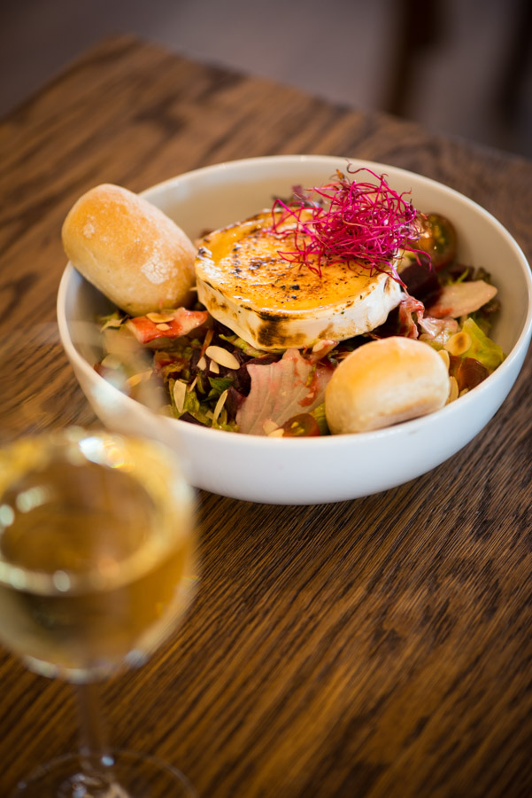 seehaus-langenfeld-salat-wein-nahaufnahme Nahaufnahme eines Ziegenkäsesalats auf einem Holztisch mit einem Weißweinglas.