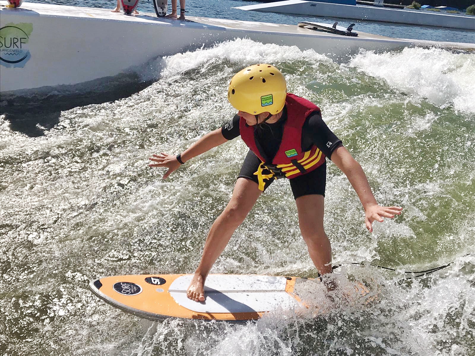 surf-langenfeld-anfänger-junior-surf-week-schulklasse Mädchen surft mit kurzem Neoprenanzug, Helm und Schwimmweste auf einem Surfboard in der stehenden Welle