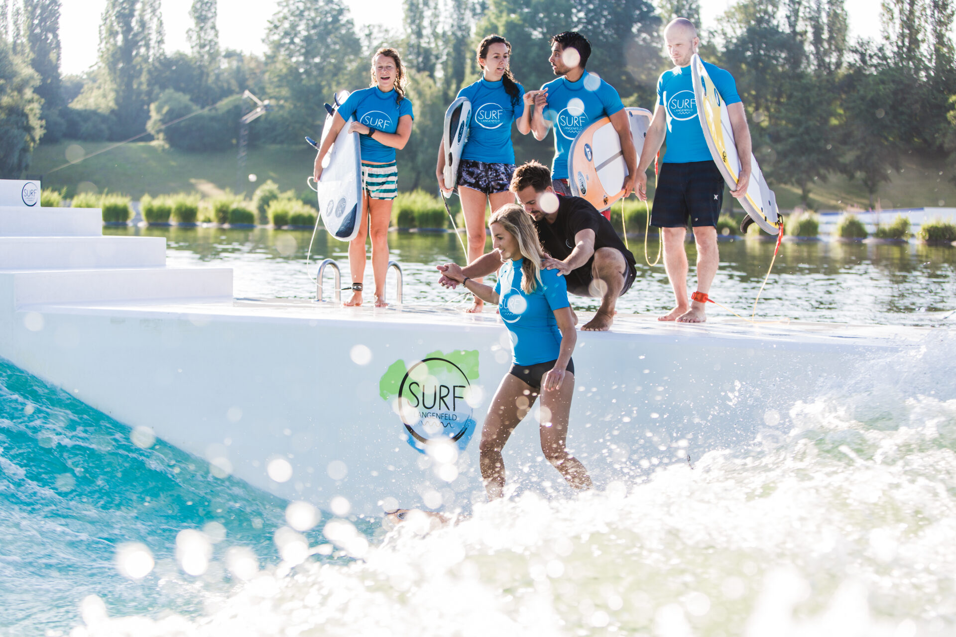 Anfängergruppe steht am Rand der stehenden Welle und beobachtet, wie ein Coach einer Anfängerin beim surfen hilft.
