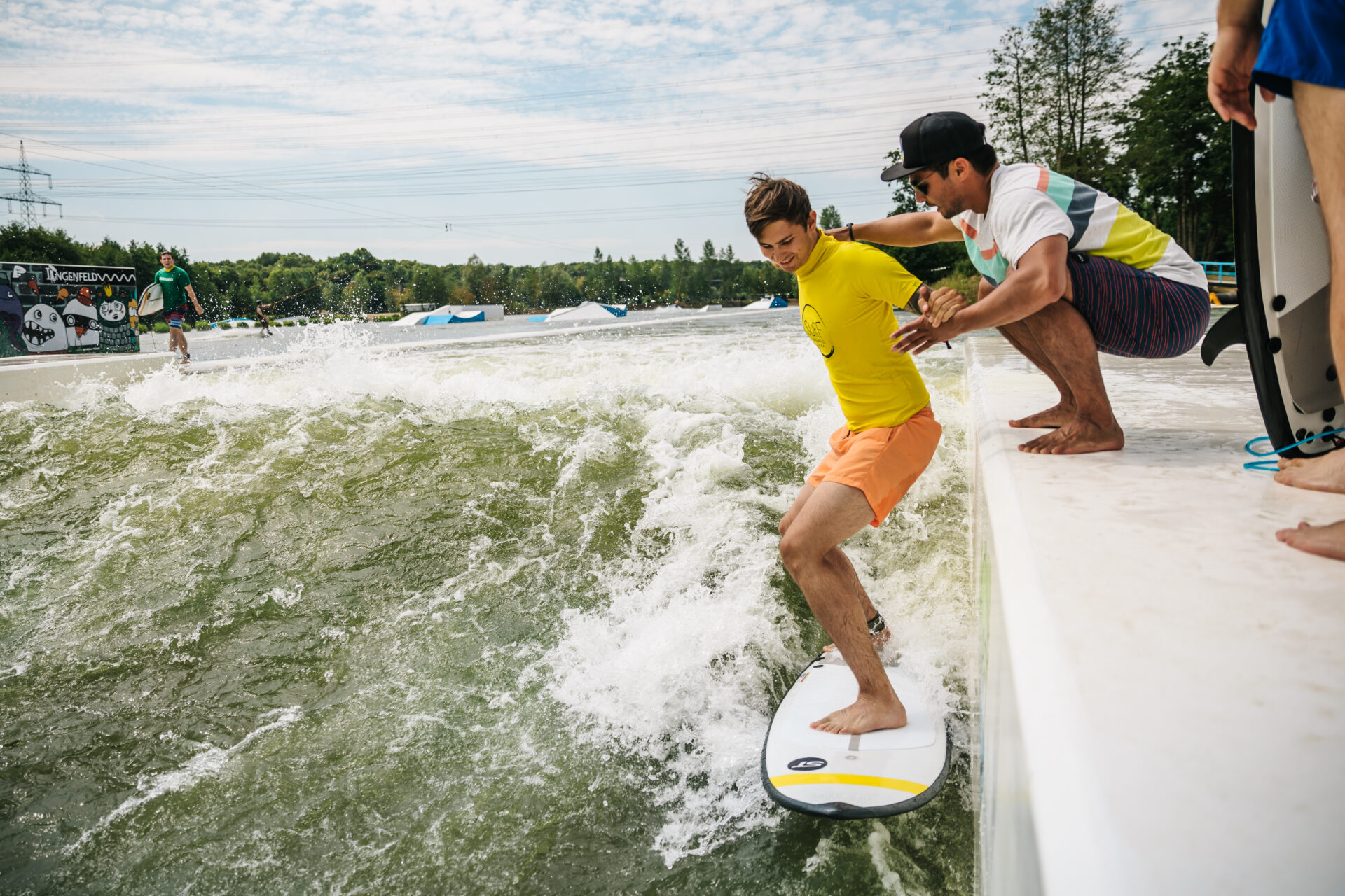 Surfcoach hocht auf dem Beckenrand der Welle und gibt einem Anfänger Hilfestellung, um in der Welle zu surfen.