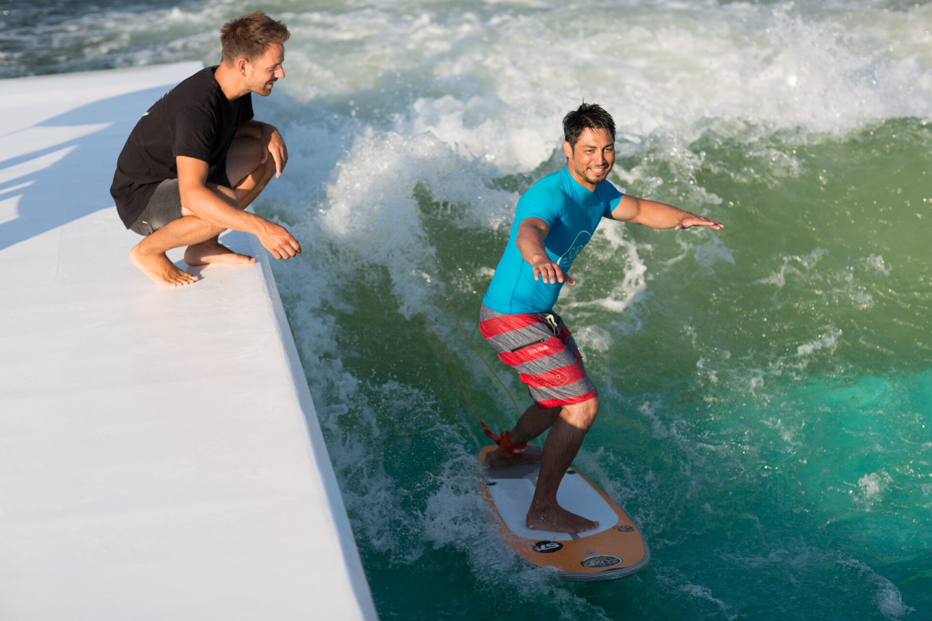 Mann surft in Anfängerhaltung auf der stehenden Welle, ein Coach hockt auf dem Beckenrand daneben und gibt Tipps.
