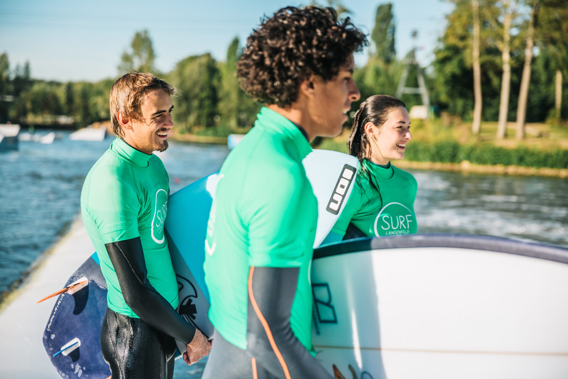 surf-langenfeld-zuschauen-surfen Drei Surfer mit grünen Lycras stehen am Beckenrand der Welle und schauen lachend auf die Welle