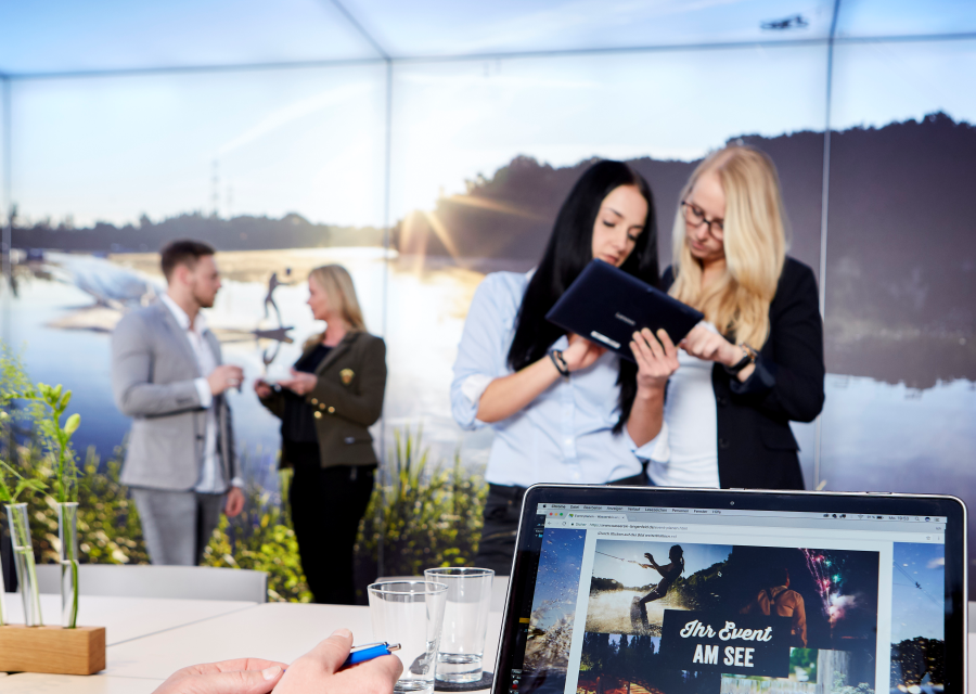 Tagungsraum. Mann und Frau schauen im Vordergrund auf einen Laptop, im Hintergrund besprechen sich ebenfalls zwei Teams