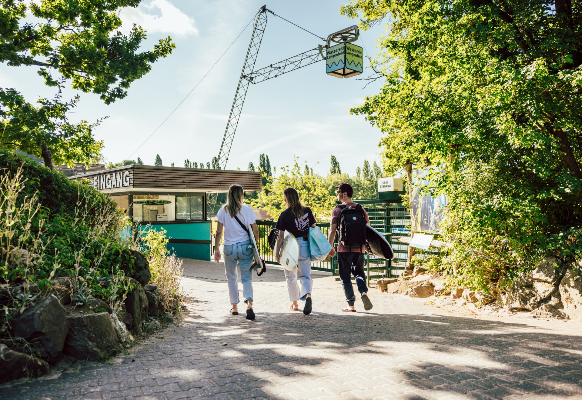 Drei Personen mit Boards unter dem Arm laufen auf den Haupteingang von Wasserski Langenfeld zu. Über ihnen ist der markante Wasserski-Langenfeld-Würfel zu sehen.